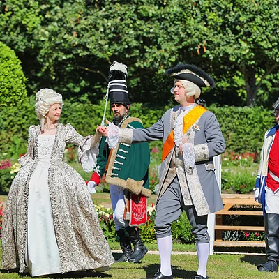 Vier Personen in historischen Kostümen aus dem Rokoko im Garten, zwei tanzen Händchen haltend.