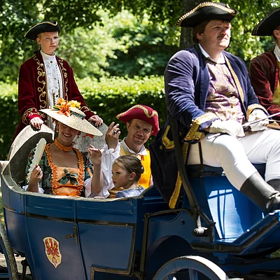 Sechs Personen in historischen Kostümen sitzen in einer blauen Kutsche im Freien unter Bäumen.