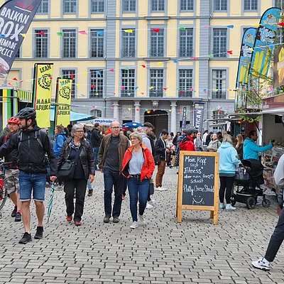 Menschen gehen auf einem gepflasterten Platz mit Essensständen und bunten Wimpelketten in einer Stadt.