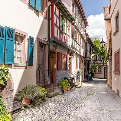 Enge gepflasterte Gasse mit Fachwerkhäusern und blauen Fensterläden bei Sonnenschein.