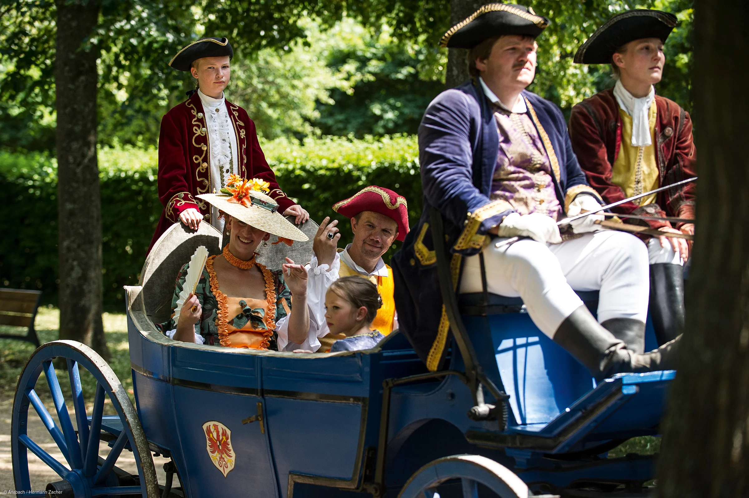 Sechs Personen in historischen Kostümen sitzen in einer blauen Kutsche im Freien unter Bäumen.