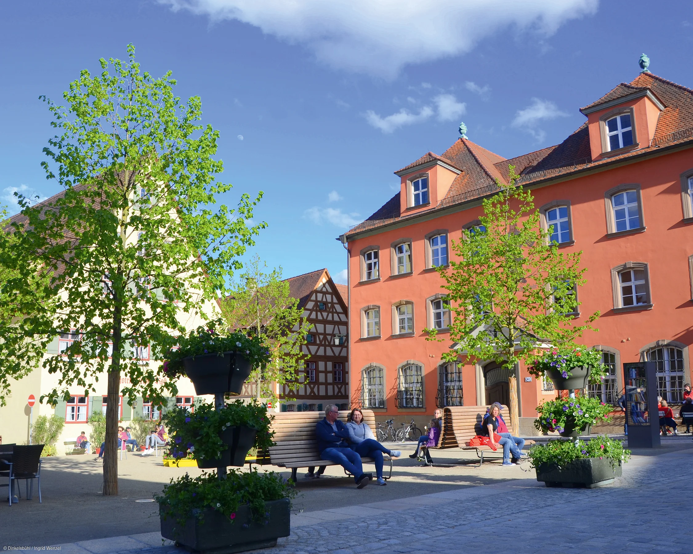 Stadtplatz mit Bänken, Menschen, Bäumen und historischen Fachwerkhäusern bei sonnigem Wetter.