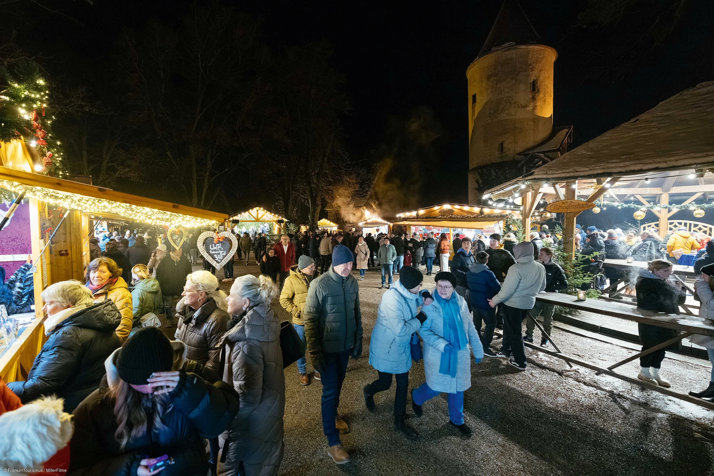 Menschen bei Nacht auf einem Weihnachtsmarkt mit Ständen und beleuchtetem Turm im Hintergrund