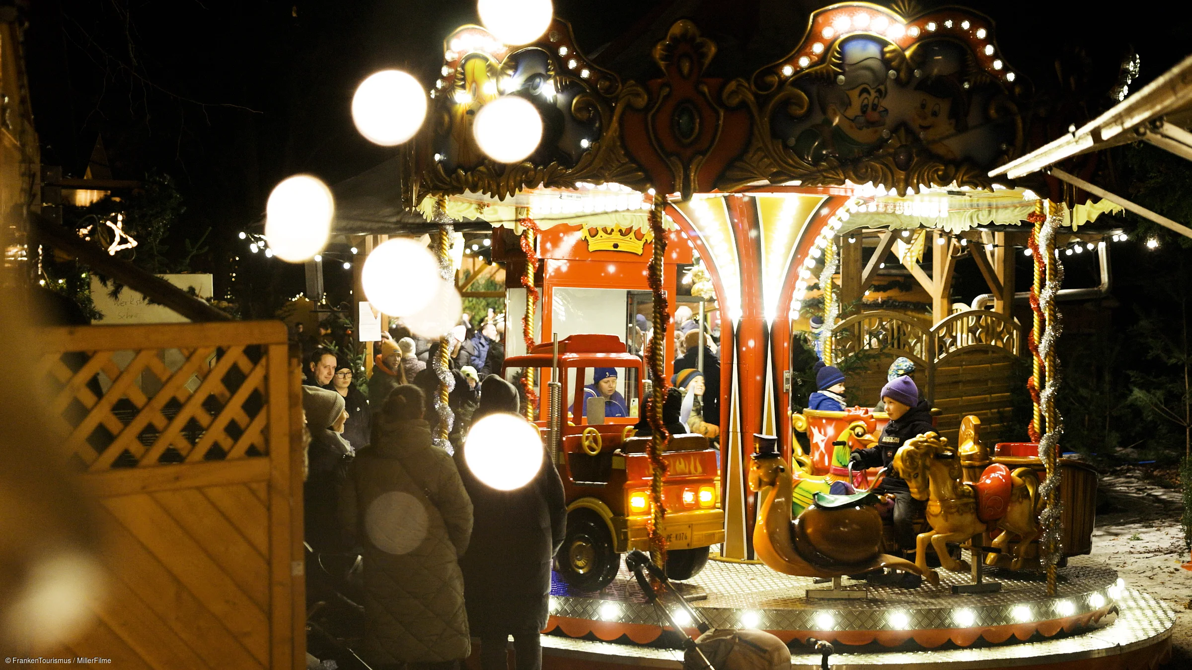Kinder fahren abends auf einem beleuchteten Karussell, umgeben von wartenden Erwachsenen.