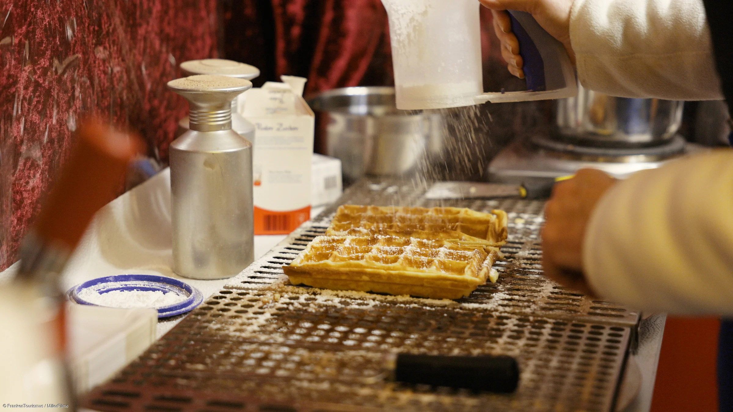 Hand streut Puderzucker auf zwei frische Waffeln auf einem Waffeleisen in einer Küche.