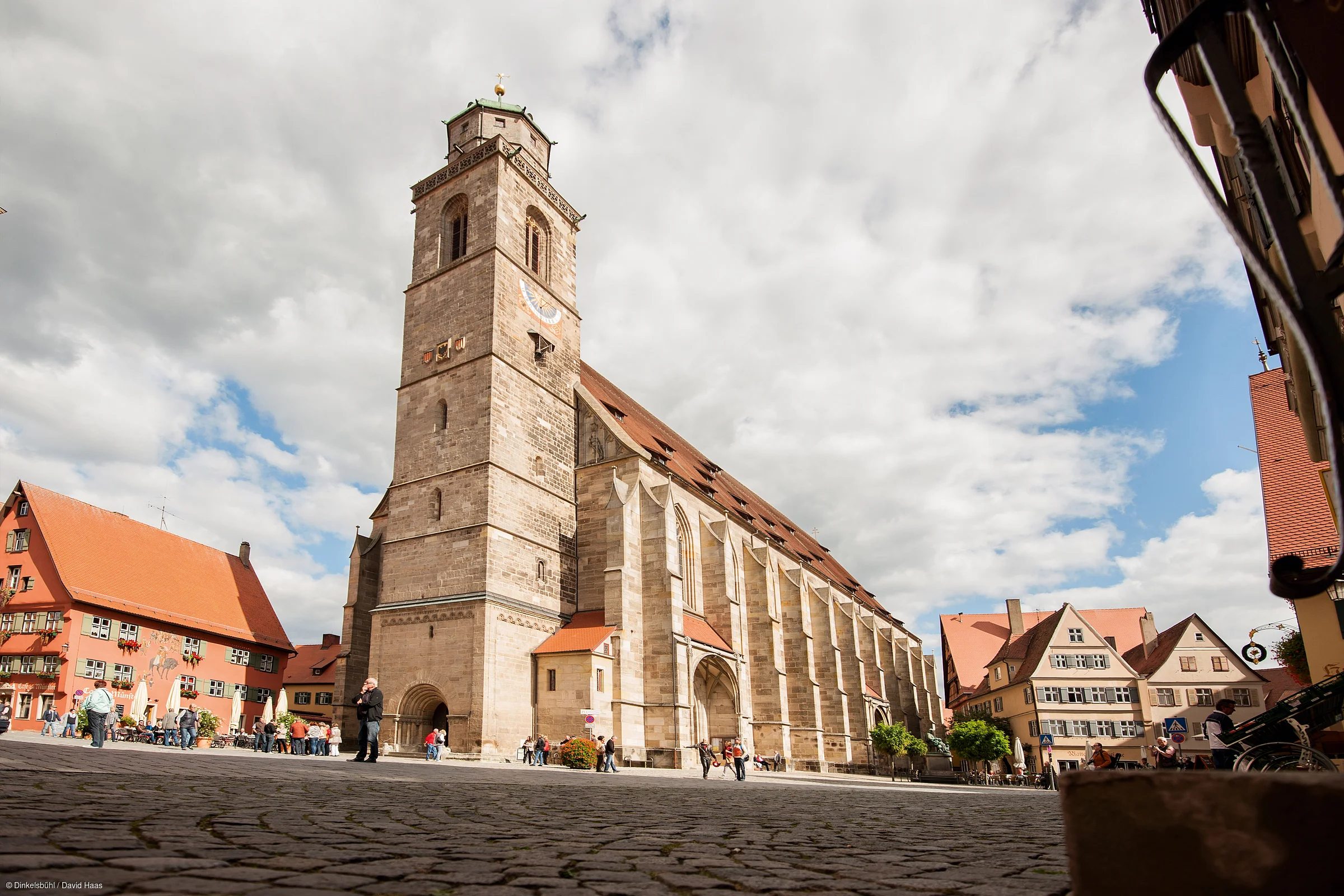 Große Kirche mit Turm auf gepflastertem Platz, umgeben von Fachwerkhäusern und Menschen bei bewölktem Himmel.