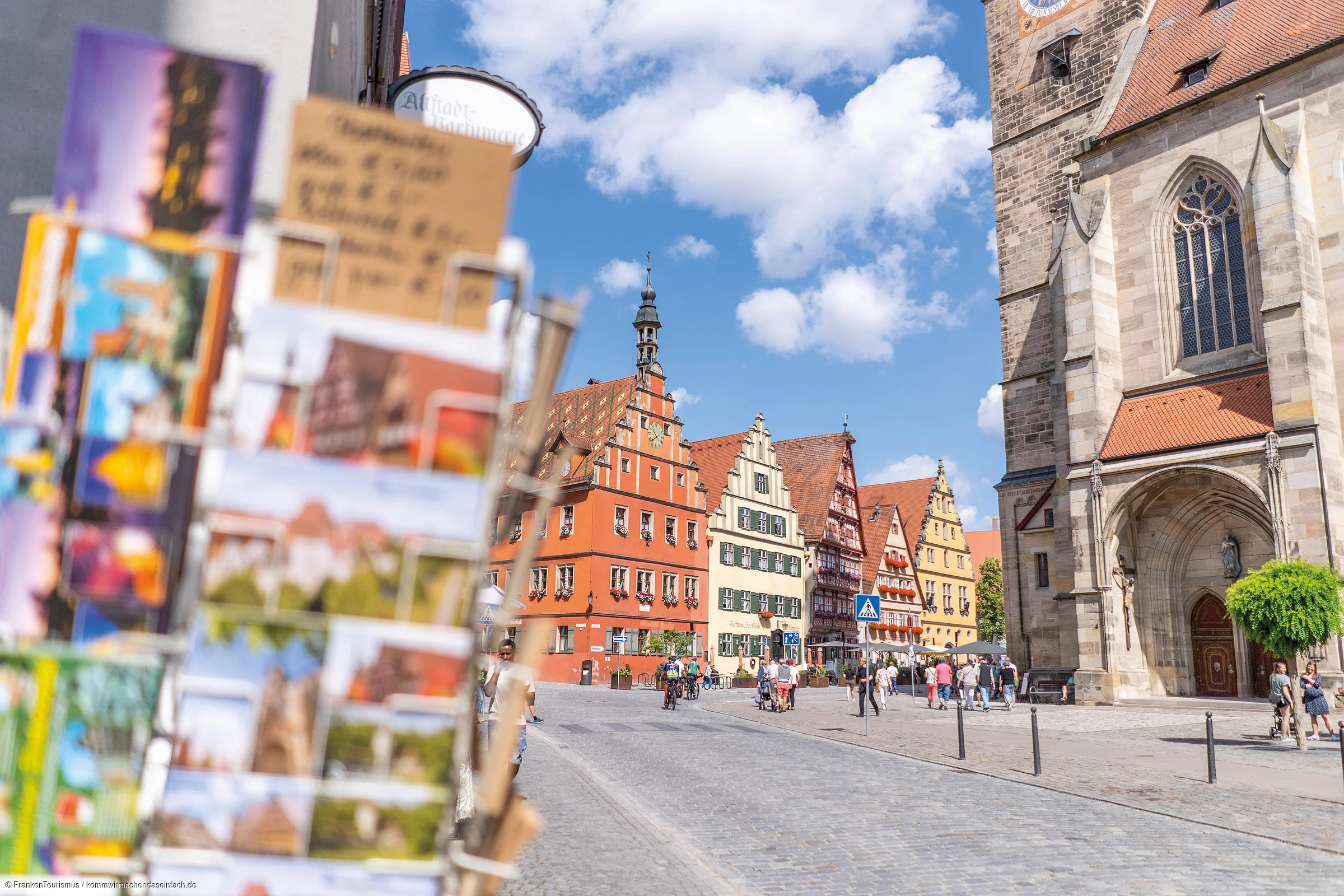 Blick auf historischen Marktplatz mit Fachwerkhäusern und Kirche bei blauem Himmel, Postkartenständer im Vordergrund unscharf.