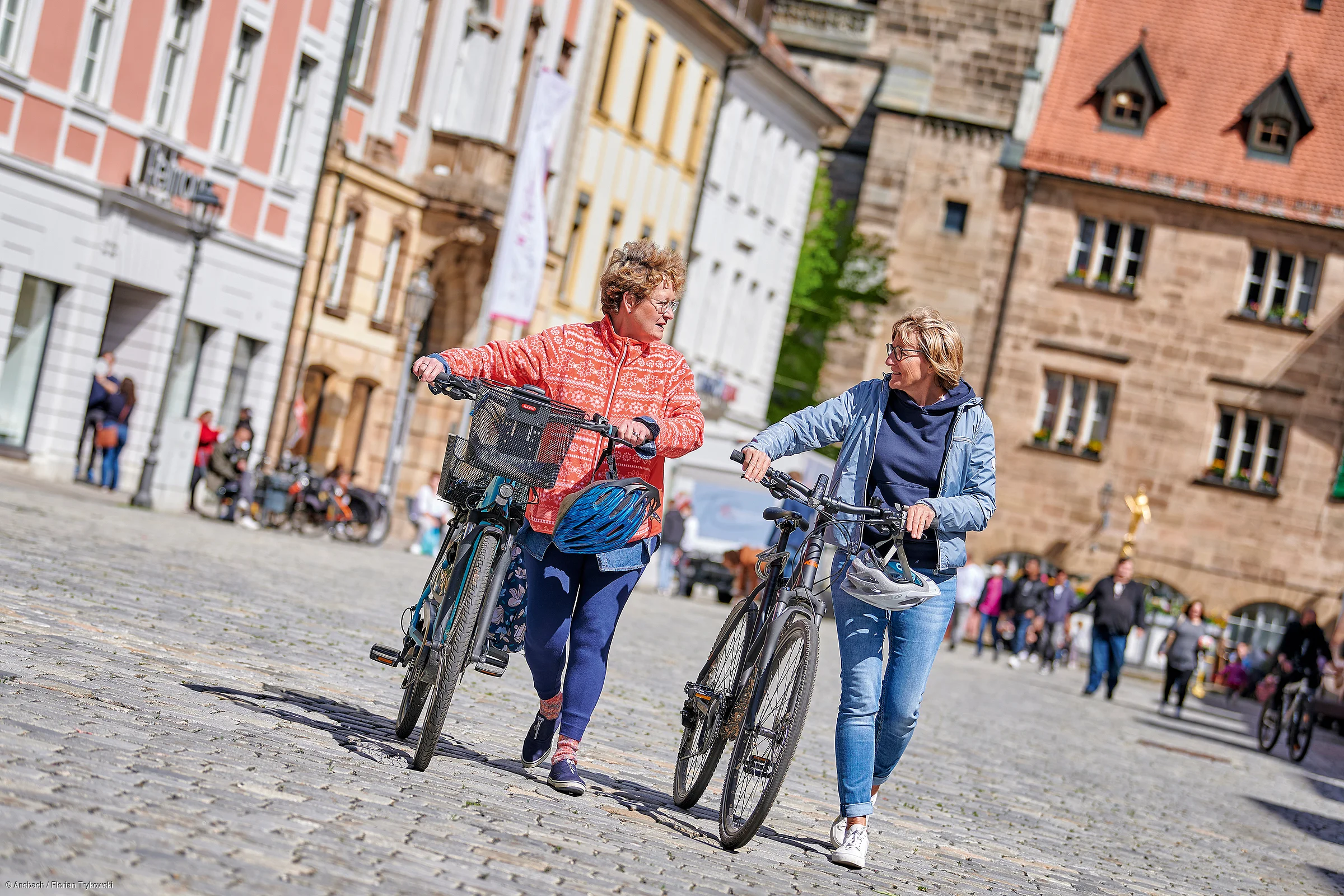 Zwei Frauen schieben Fahrräder auf einem gepflasterten Platz vor historischen Gebäuden.