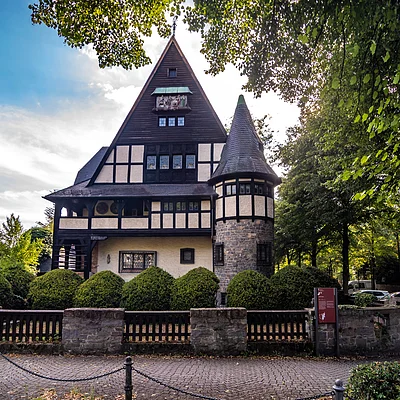 Fachwerkhaus mit spitzem Giebel und rundem Turm, umgeben von Büschen und Bäumen an einem gepflasterten Weg.