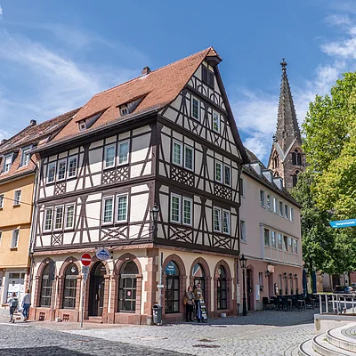 Fachwerkhaus an einer Straßenecke mit Kopfsteinpflaster und Kirchturm im Hintergrund bei blauem Himmel.