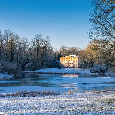 Schloss am zugefrorenen See mit schneebedecktem Boden und kahlen Bäumen im Winter bei klarem Himmel.