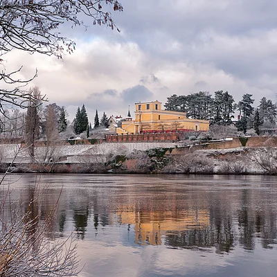 Gelbes Gebäude auf einem schneebedeckten Hügel mit Bäumen, Spiegelung im ruhigen Fluss im Vordergrund.