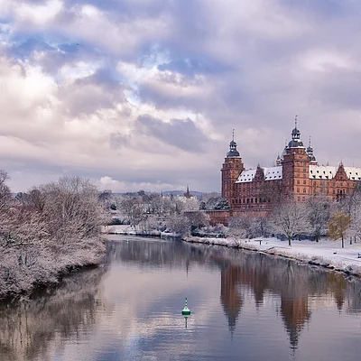 Winterliche Flusslandschaft mit schneebedeckten Bäumen und einem historischen Schloss im Hintergrund