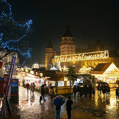 Weihnachtsmarkt bei Nacht mit beleuchteten Ständen, Menschen mit Regenschirmen und Schloss im Hintergrund.