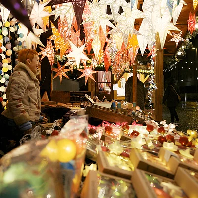 Weihnachtsmarktstand mit leuchtenden Papiersternen und Kunden bei Nacht in Winterkleidung