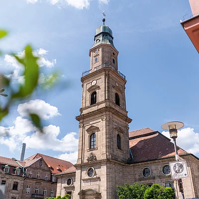 Kirchturm eines historischen Gebäudes bei blauem Himmel, unscharfe Blätter im Vordergrund, Straßenschild sichtbar