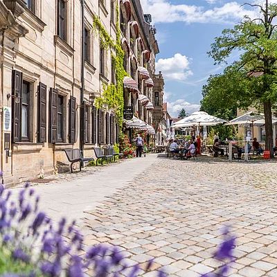 Pflasterplatz mit Sitzgruppen und Sonnenschirmen vor historischen Gebäuden und Bäumen an einem sonnigen Tag