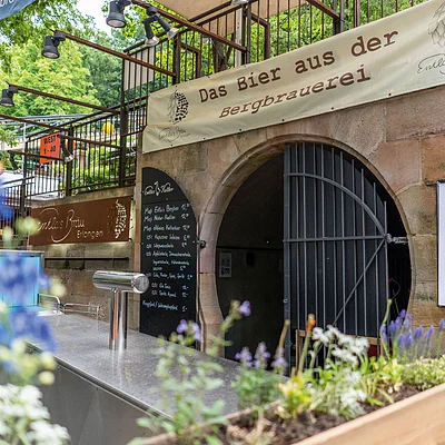 Außenbereich einer Brauerei mit Zapfhahn, Blumen im Vordergrund und Schild „Das Bier aus der Bergbrauerei“.