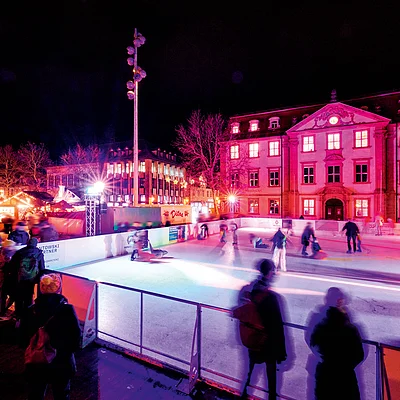 Eisbahn mit mehreren Eisläufern und beleuchteten Gebäuden bei Nacht in einer Stadt.