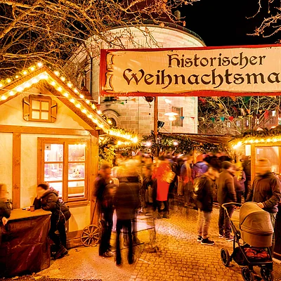 Menschen auf historischem Weihnachtsmarkt mit beleuchteten Buden und Banner bei Nacht.