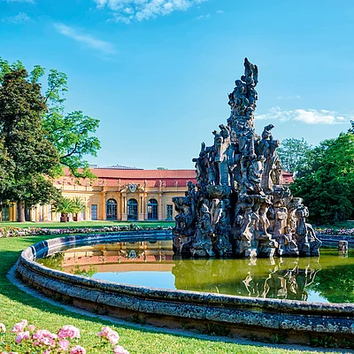 Brunnen mit Figuren in einem runden Wasserbecken vor einem gelben Schloss und blühenden Rosen im Park bei blauem Himmel