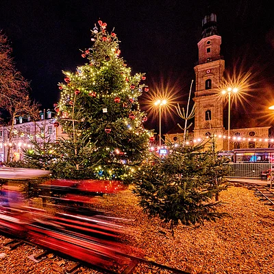 Weihnachtsmarkt bei Nacht mit beleuchtetem Weihnachtsbaum, Mini-Zug und historischem Turm im Hintergrund.