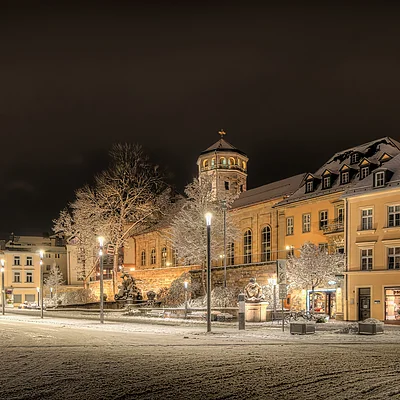 Schneebedeckte Straße mit beleuchteten Gebäuden und Bäumen bei Nacht in einer Stadt.