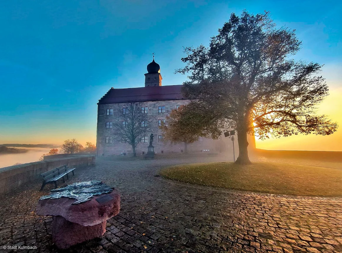 Schloss mit Turm bei Sonnenaufgang, Baum rechts, Bank und Steinplatte links auf gepflastertem Platz