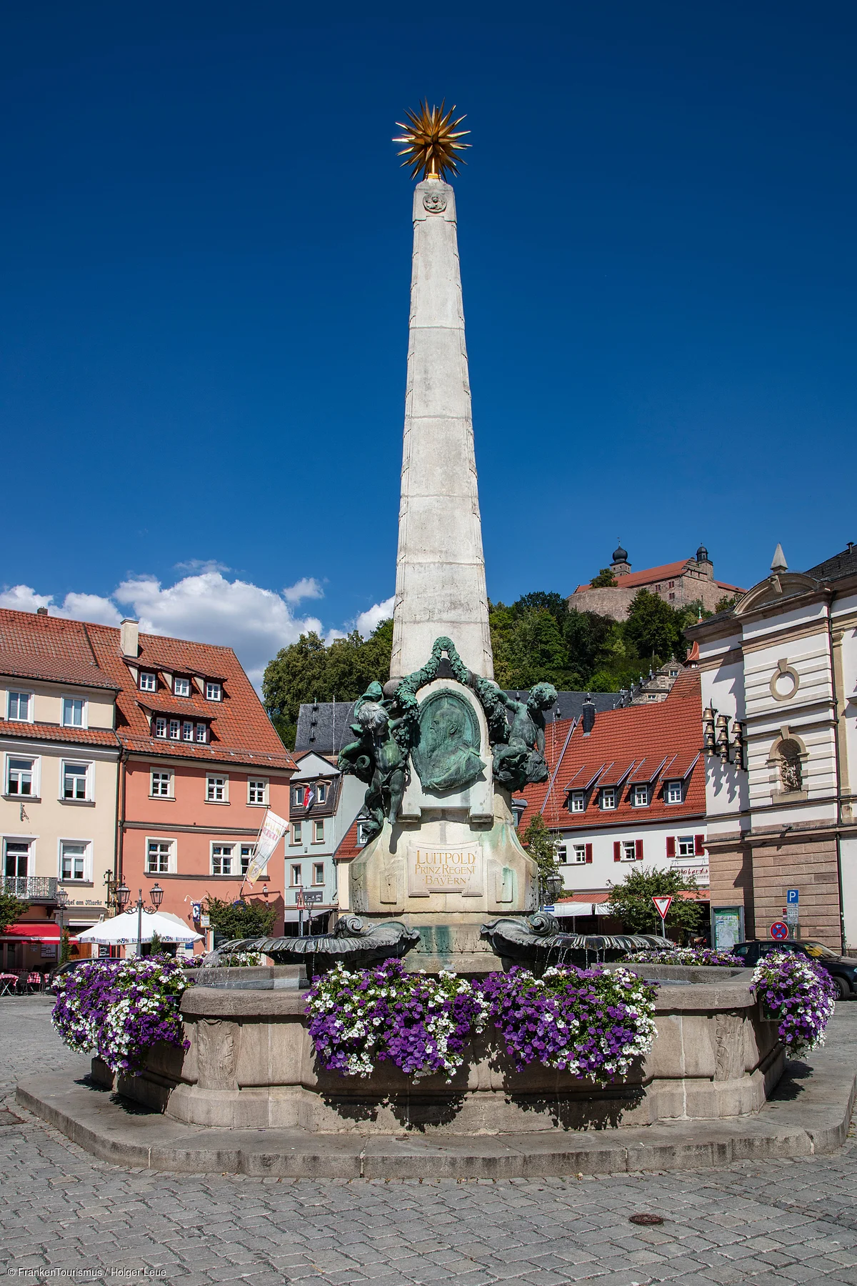 Obelisk mit bronzenem Relief und goldener Sternspitze auf gepflastertem Platz mit Blumen und Häusern im Hintergrund