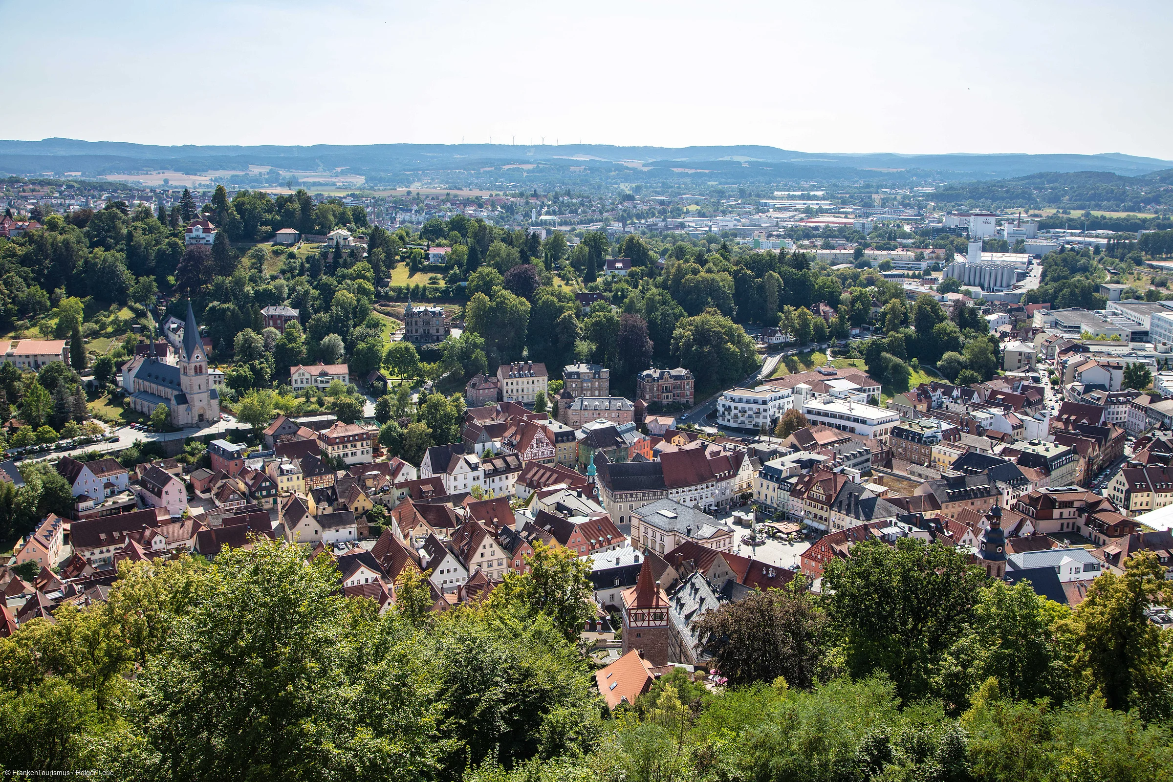 Blick auf eine Stadt mit vielen Häusern, Bäumen und Hügeln im Hintergrund bei klarem Himmel.
