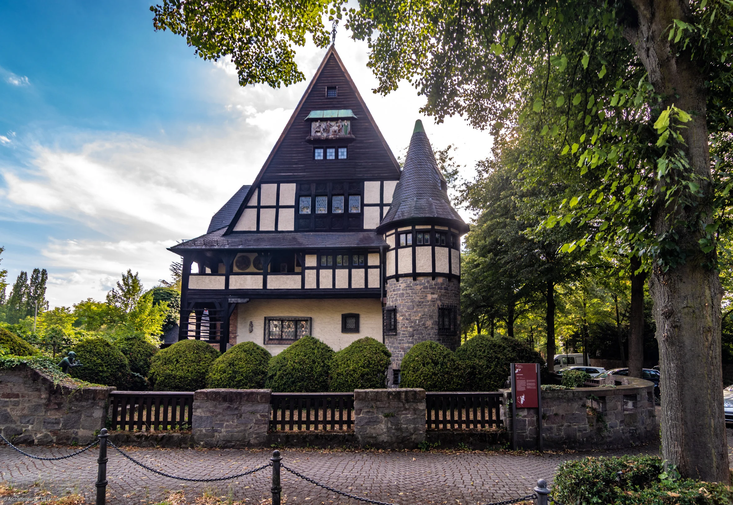 Fachwerkhaus mit spitzem Giebel und rundem Turm, umgeben von Büschen und Bäumen an einem gepflasterten Weg.