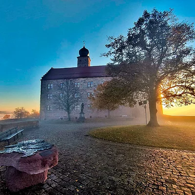 Schloss mit Turm bei Sonnenaufgang, Baum rechts, Bank und Steinplatte links auf gepflastertem Platz