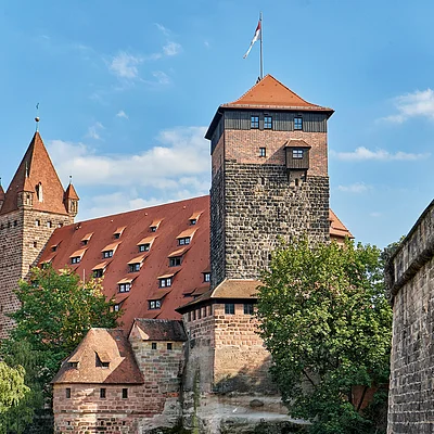 Burg mit roten Dächern und Türmen, umgeben von Bäumen und einer Steinmauer bei blauem Himmel.