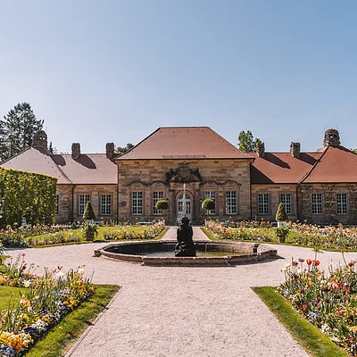 Barocker Garten mit Blumenbeeten, Springbrunnen und einem historischen Gebäude im Hintergrund bei klarem Himmel.