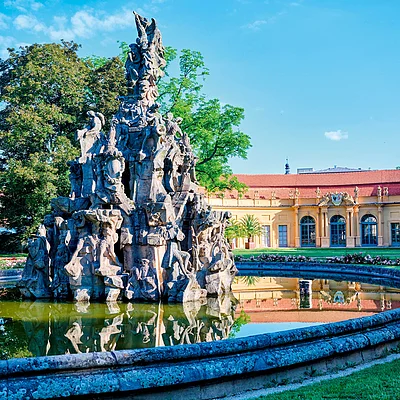 Steinerne Skulptur mit Figuren in rundem Wasserbecken vor gelbem Gebäude und Bäumen bei blauem Himmel.
