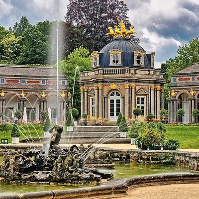 Barockgarten mit Springbrunnen und Pavillon mit goldener Quadriga auf dem Dach, umgeben von Bäumen und Rasenflächen