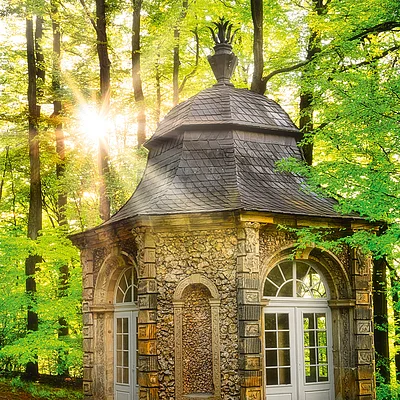 Kleines steinernes Pavillongebäude mit weißen Türen im Wald bei Sonnenlicht am Morgen.