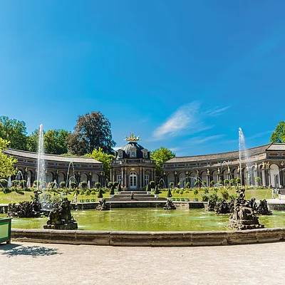 Brunnen mit Wasserspielen vor einem historischen Gebäude und Bäumen unter blauem Himmel.