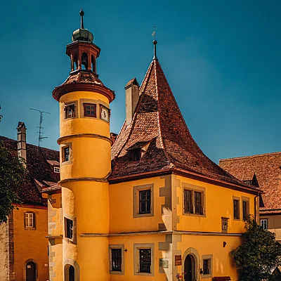 Gelbes historisches Gebäude mit Turm und spitzem Dach bei sonnigem Himmel auf gepflastertem Platz.