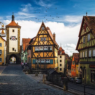 Altstadt mit Fachwerkhäusern, Kopfsteinpflasterstraße und einem Turm mit Uhr unter blauem Himmel mit Vögeln