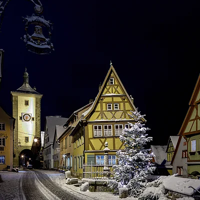 Schneebedeckte Fachwerkhäuser und ein beleuchteter Turm bei Nacht in einer historischen Altstadt.