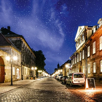 Pflasterstraße mit parkenden Autos und beleuchteten historischen Gebäuden unter sternklarem Nachthimmel.