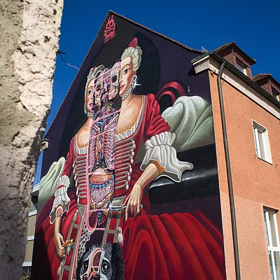 Wandgemälde einer historischen Frau mit teilweise sichtbaren inneren Organen an der Fassade eines Gebäudes.