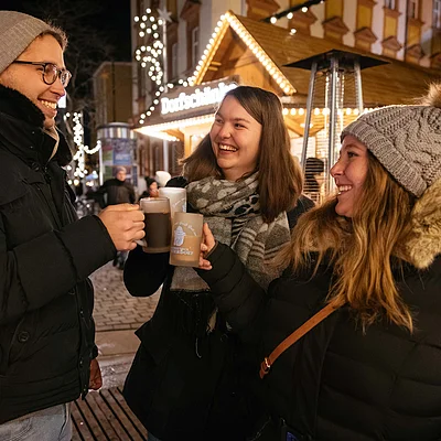 Drei Personen stoßen mit Tassen bei einem beleuchteten Weihnachtsmarktstand im Freien an.