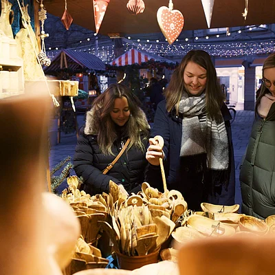 Drei Frauen in Winterjacken betrachten Holzlöffel und -schalen an einem Marktstand mit Weihnachtsdekoration.