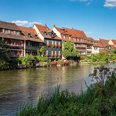 Flussufer mit grüner Vegetation und Fachwerkhäusern mit roten Dächern unter blauem Himmel am Wasser.