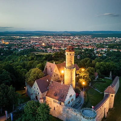 Luftaufnahme einer beleuchteten Burg mit Stadt und Wald im Hintergrund bei Abenddämmerung.