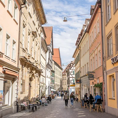 Fußgängerzone mit Cafés, Menschen und historischen Gebäuden unter blauem Himmel mit Wolken.