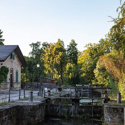 Altes Schleusentor mit Wasser, umgeben von Bäumen und einem Gebäude mit grünem Fensterladen.