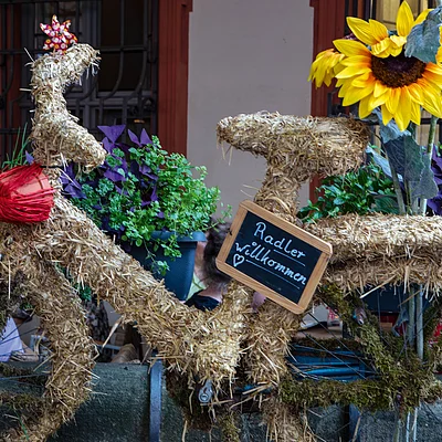 Fahrrad komplett mit Stroh bedeckt, mit Sonnenblumen und Schild „Radler willkommen“ am Rahmen.